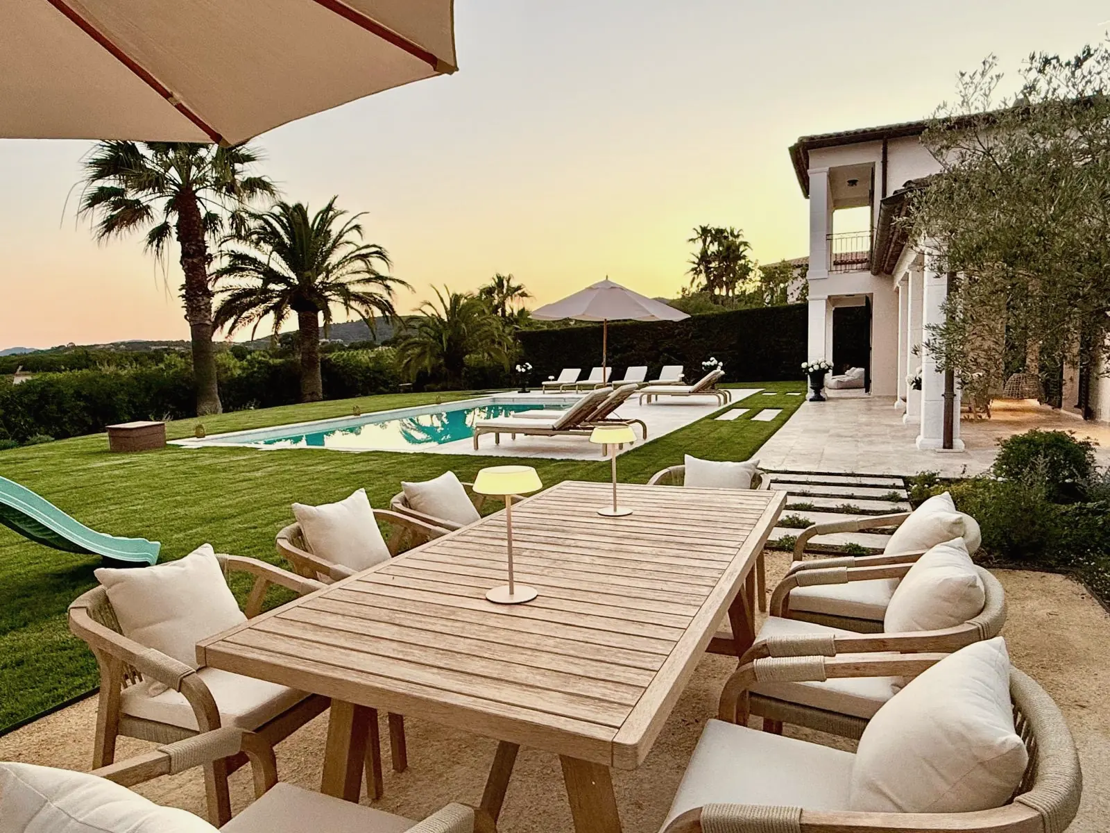 Mesa de teca bajo una sombrilla en la terraza al atardecer, con la piscina detrás